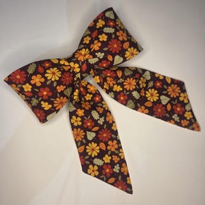 May include: A fabric bow with a fall floral pattern. The bow features a dark brown background with orange, yellow, and red flowers, along with green leaves. The bow is tied in the center and has long, pointed tails.