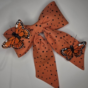 Puede incluir: Un lazo de tela naranja con lunares negros, adornado con dos mariposas monarca decorativas. Las mariposas tienen alas naranjas y negras con puntos blancos. Accesorio decorativo.