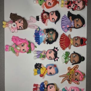 May include: A collection of colorful doll-shaped embellishments, each with unique outfits and hairstyles. The dolls feature various designs, including a fairy, a strawberry, and a character with a black cat. These decorative pieces are ideal for crafting and scrapbooking projects.