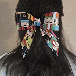 Op de afbeelding: Een kleurrijke haarstrik met een herhaald patroon van notenkrakerfiguren in verschillende uniformen. De strik is in een klassieke stijl geknoopt en staat tegen een achtergrond van donker haar. Het ontwerp van de strik bevat rode, blauwe en gele accenten.