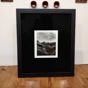 May include: A black framed photograph of a rocky landscape with a cloudy sky. The photograph is in a black frame with a white mat.