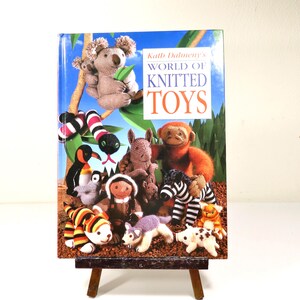 May include: A hardcover book titled Kath Dalmeny's World of Knitted Toys. The cover features a colorful collection of various knitted animal figures, including a koala, a monkey, a zebra, a penguin, and a snake, set against a blue sky background.