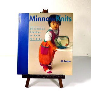 May include: A softcover book titled MinnowKnits: Uncommon Clothes to Knit for Kids by Jil Eaton. The cover features a child wearing a bright orange knitted garment with a pink pocket, standing next to a glass fishbowl containing a goldfish.