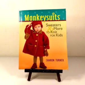 May include: A softcover book titled Monkeysuits: Sweaters & More to Knit for Kids by Sharon Turner. The cover features a child wearing a red knitted coat with a matching hat and shoes, set against a teal and orange background with white text.