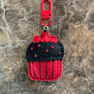 May include: A red and black felt cupcake keychain with a red clasp. The cupcake has a red base and a black top with red dot accents. The keychain is designed to resemble a miniature cupcake.