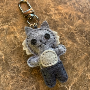 May include: A gray felt keychain shaped like a small creature with a round light gray belly, dark gray pants, and black circular eyes. The figure features pointed ears and a silver-toned metal lobster clasp attached to the top with a ring.