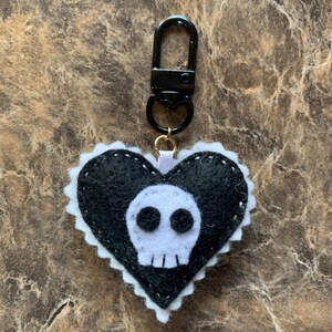 Op de afbeelding: Een zwarte vilten hartvormige sleutelhanger met een wit schedelontwerp. Het hart heeft een witte geschulpte rand en is bevestigd aan een zwarte metalen sluiting. Een accessoire met een gotisch thema.