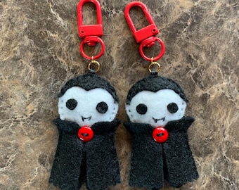 May include: Two felt vampire keychains featuring white faces with black circular eyes and small fangs, wearing black capes fastened with a single red button. Each figure is attached to a bright red plastic swivel lobster clasp hook.