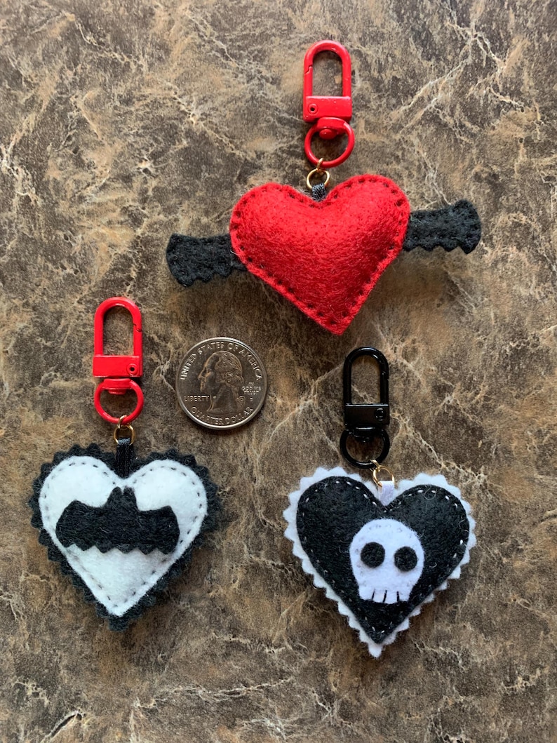 Op de afbeelding: Drie hartvormige sleutelhangers. Een rood hart met zwarte vleugels, een zwart-wit hart met een vleermuis en een zwart-wit hart met een schedel. Elke sleutelhanger heeft een rode of zwarte sluiting.