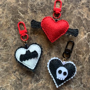 Op de afbeelding: Drie hartvormige sleutelhangers. Een rood hart met zwarte vleugels, een zwart-wit hart met een vleermuis en een zwart-wit hart met een schedel. Elke sleutelhanger heeft een metalen sluiting.