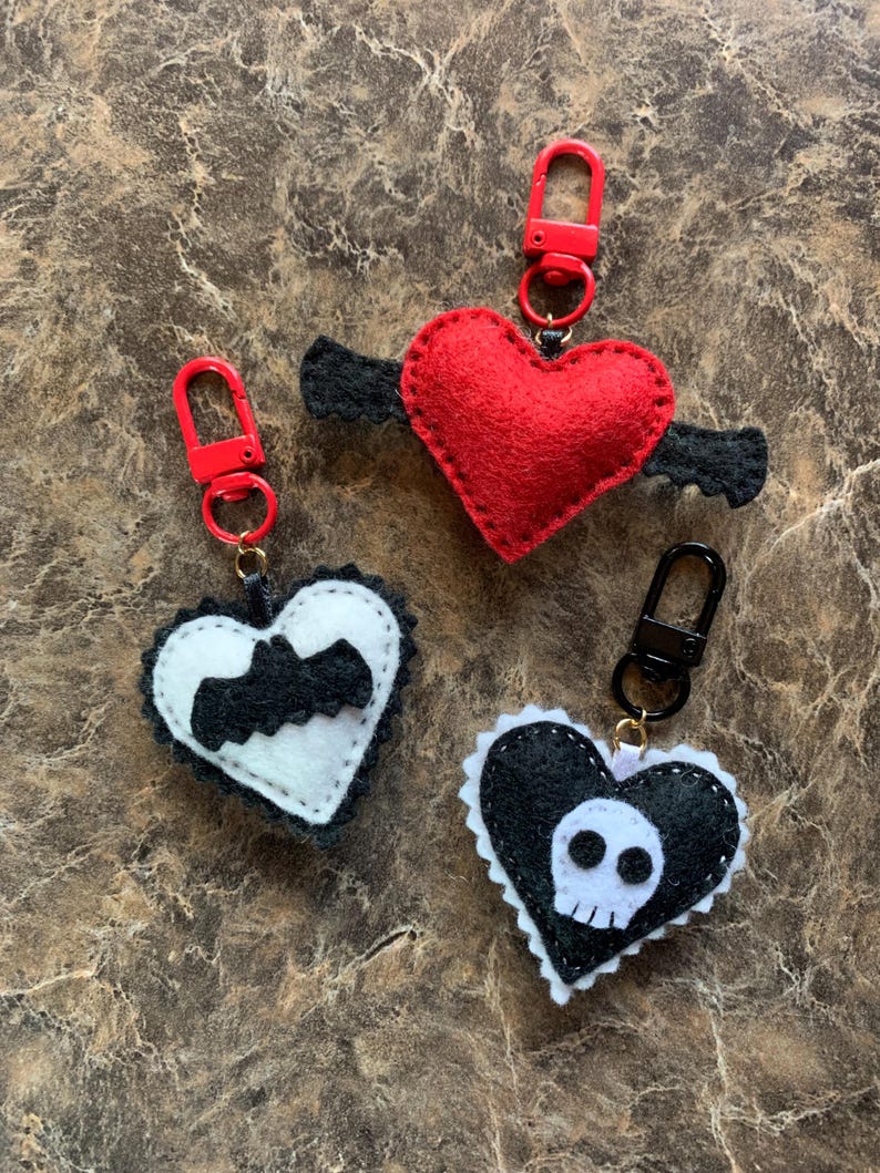 Op de afbeelding: Drie hartvormige sleutelhangers. Een rood hart met zwarte vleugels, een zwart-wit hart met een vleermuis en een zwart-wit hart met een schedel. Elke sleutelhanger heeft een metalen sluiting.