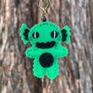 May include: A handmade green felt keychain featuring a whimsical monster design. The monster has black accents, including a circular belly, a smiling mouth, and two eyes. The keychain is attached to a gold-colored metal clasp.