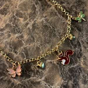 May include: Gold-toned charm bracelet featuring a butterfly, squirrel, dragonfly, and heart charms. The butterfly is pink, the squirrel is brown and white, and the dragonfly is green. The bracelet is on a marble surface.