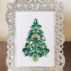 May include: A decorative silver picture frame with a sparkling Christmas tree design. The tree is made of various sizes of green and light green gemstones. The frame has an ornate, detailed border with clear crystals.