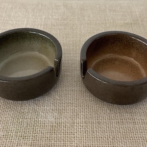 May include: Two ceramic ashtrays with a dark brown exterior and a speckled interior. One ashtray has a green interior, and the other has a brown interior. Each has a curved design with a notch for resting items.