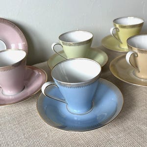 Op de afbeelding: Een set van zes pastelkleurige theekopjes en schotels. De kopjes en schotels zijn in tinten roze, geel, groen en blauw, met gouden randen. De kopjes hebben een klassiek ontwerp en de schotels zijn rond.