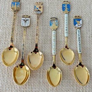 May include: Set of seven gold-tone souvenir spoons with enamel city crests and names. The spoons feature crests from the cities of Lillehammer, Drammen, Oslo, Holmen, Drammen, and Stonsberg.