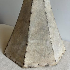 May include: A handmade lampshade with a unique, geometric shape. The shade is constructed from a light-colored, textured material, possibly leather or a similar fabric. It features visible stitching along the seams and edges, adding to its rustic charm. The shade is designed to fit over a lamp base.