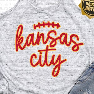 May include: A gray tank top with a red and yellow graphic that says "kansas city" with a football design above the text. The tank top is laying on a white surface with a pair of white sneakers and a pair of blue denim shorts.