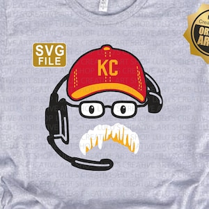 May include: A gray t-shirt with a cartoon illustration of a person wearing a red and white baseball cap with the letters "KC" on it. The person is wearing glasses and a mustache. The illustration is surrounded by a black circle and a headset.