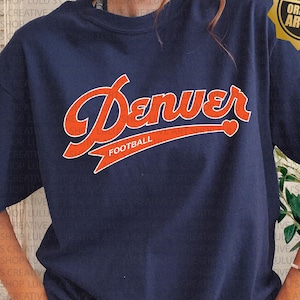 May include: Navy blue t-shirt with the word "Denver" in orange script, and "FOOTBALL" below. The shirt has a classic crew neck and short sleeves. A gold seal that says "Lulu's Creative ArtShop ORIGINAL ARTIST" is in the upper right corner.