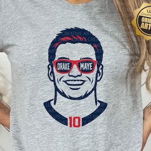 May include: A light gray t-shirt featuring a graphic of a smiling person wearing red sunglasses with the text "DRAKE MAYE". The number "10" is below the graphic. The shirt also has the text "SVG/PNG".