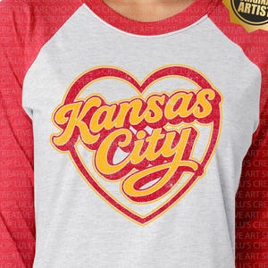 May include: A raglan baseball tee with a white body and red sleeves. The shirt features a distressed heart-shaped graphic with the words "Kansas City" in yellow and red. The text "PNG (distressed)" is at the bottom.
