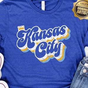May include: A blue t-shirt with a retro-style graphic that says "Kansas City" in white, yellow, and blue lettering. The graphic has a distressed look and is surrounded by a white, yellow, and blue wavy design. The t-shirt is laid flat on a wooden surface with a pair of white sneakers and a pair of blue denim shorts.