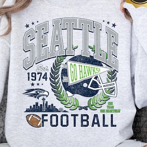May include: A light grey sweatshirt with a distressed graphic design. The design features the word "SEATTLE" above "FOOTBALL", a football helmet, and the text "GO HAWKS!". The sweatshirt also includes the text "Est. 1974".
