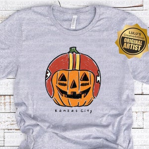 May include: A light gray t-shirt featuring a pumpkin wearing a football helmet. The pumpkin has a carved face and the text "Kansas City" is below the pumpkin graphic. The shirt has a crew neck.