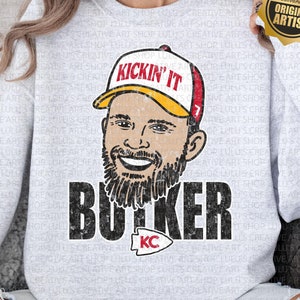May include: A gray sweatshirt with a distressed graphic of a football player wearing a red and yellow hat with the text "Kickin' It" and the name "Butker" with a red and yellow KC logo. The sweatshirt has a distressed look and is a PNG file.