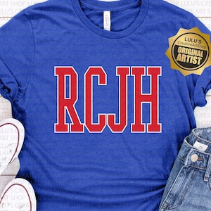 Puede incluir: Una camiseta azul con las letras RCJH en rojo en la parte delantera. La camiseta está sobre una superficie de madera con un par de zapatillas blancas y unos pantalones cortos de mezclilla azul. Una pegatina dorada con el texto "Lulu's Creative ArtShop Original Artist" está en la camiseta.
