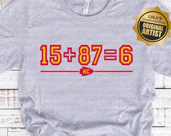 May include: A gray t-shirt with a red and yellow graphic that says "15+87=6" with a red arrow pointing to the right and the letters "KC" underneath. The graphic is on a gray t-shirt.