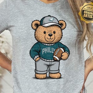 Op de afbeelding: Een grijs gemêleerd t-shirt met een bruine teddybeer-afbeelding die een groene pet met adelaarslogo, een groene trui met de tekst PHILLY en een grijze broek draagt terwijl hij een bruine football vasthoudt. Het shirt heeft een gouden zegel met LULU's CREATIVE ART SHOP ORIGINAL ARTIST.
