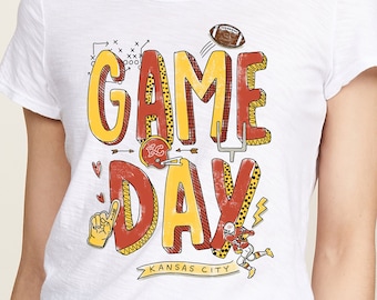 May include: White t-shirt with a red and yellow graphic that says "GAME DAY" with a football, a football helmet, and a football player. The text "KANSAS CITY" is below the graphic.