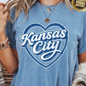 May include: Light blue t-shirt featuring a white and blue heart-shaped design with the words "Kansas City" in a script font. The shirt has short sleeves and a distressed look. A "Original Artist" sticker is visible.