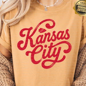 May include: A mustard yellow short-sleeve t-shirt featuring the red text Kansas City in a cursive script font with a small arrowhead icon above the word Kansas. The shirt has a crew neckline and is worn with a brown knitted cardigan.