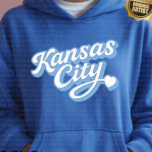May include: Blue hoodie with the text "Kansas City" in a white and light blue design. The hoodie has a drawstring and a front pocket. The image also includes a gold sticker that says "Original Artist".