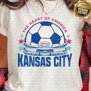 以下が含まれることがあります： クリーム色のTシャツ。デザインには、サッカーボール、スタジアム、テキスト「THE HEART OF AMERICA」と「KANSAS CITY」が含まれています。シャツには「ARROWHEAD」の文字も入っています。