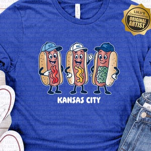 May include: Royal blue t-shirt featuring a graphic of three animated hot dogs wearing baseball caps, each with different toppings. The text "Kansas City" is printed below the hot dogs. A gold "Original Artist" seal is in the upper right corner.