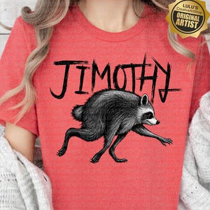 May include: A heathered red short-sleeve t-shirt featuring a black graphic illustration of a raccoon in a running pose. Above the raccoon, the text JIMOTHY is printed in a bold, distressed black font. A gold-colored badge is in the top right corner.