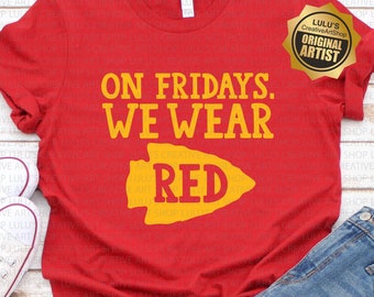 May include: Red t-shirt with the text "On Fridays We Wear Red" and a yellow arrowhead graphic. The shirt is on a wooden surface with a pair of white sneakers and blue denim shorts.