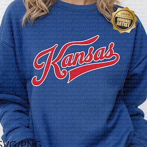May include: A blue sweatshirt with the word "Kansas" in red script lettering with a white outline. The sweatshirt also has a gold "Original Artist" seal.