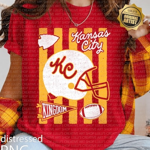May include: A red t-shirt featuring a graphic print with vertical yellow stripes. The design includes a white football helmet with the letters KC, a football, an arrowhead, a pennant with the word KINGDOM, and the text Kansas City.