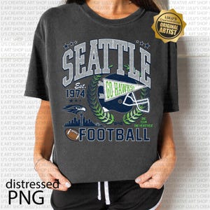 May include: Dark grey t-shirt with a distressed design featuring "SEATTLE" in large letters, a football helmet, and the text "FOOTBALL." The shirt includes the text "Est. 1974" and a logo with the words "GO HAWKS."