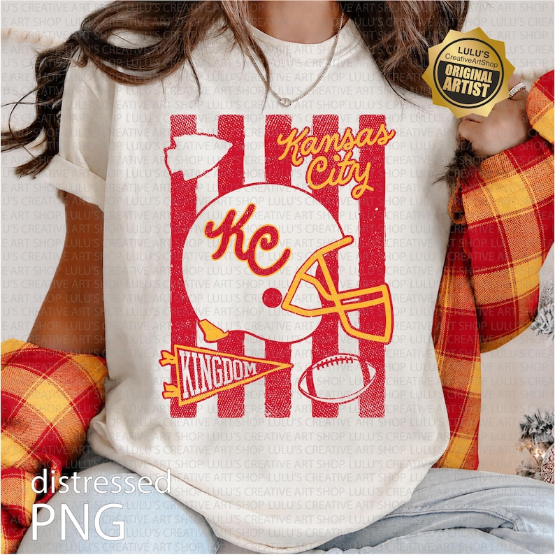 May include: A cream-colored t-shirt featuring a red and yellow graphic with vertical red stripes. The design includes a football helmet with the letters KC, a football, a pennant labeled KINGDOM, and the text Kansas City in yellow script.