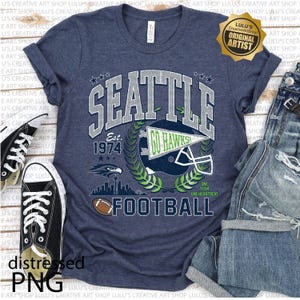 May include: Navy blue t-shirt with a distressed design featuring the word "SEATTLE" above a football helmet and the text "GO HAWKS". The shirt also includes the text "EST. 1974" and a football graphic. A pair of black trainers and jeans are next to the shirt.