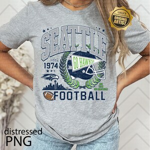 May include: A grey t-shirt with a Seattle football design. The design includes the text "SEATTLE", "GO HAWKS", a football helmet, and the year "1974". The word "FOOTBALL" is printed at the bottom.