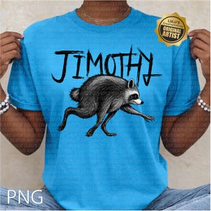 May include: A bright blue t-shirt featuring a black ink-style illustration of a raccoon in a running pose. Above the raccoon, the name JIMOTHY is printed in a bold, hand-drawn black font. A gold-colored seal in the top right corner reads ORIGINAL ARTIST.