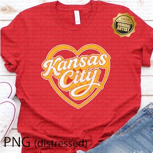 May include: Red t-shirt featuring a distressed design with the words "Kansas City" in a heart-shaped graphic. The design has a vintage look with yellow and white lettering. Includes a gold seal that says "Original Artist".